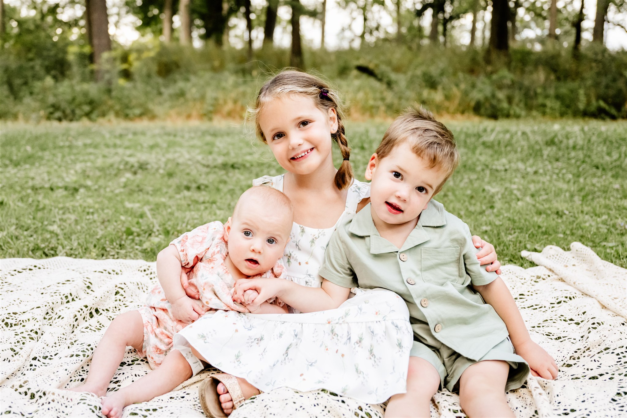 A young girl and boy hold their baby sister on a laced picnic blanket in a park after visiting splash pads in chicago.