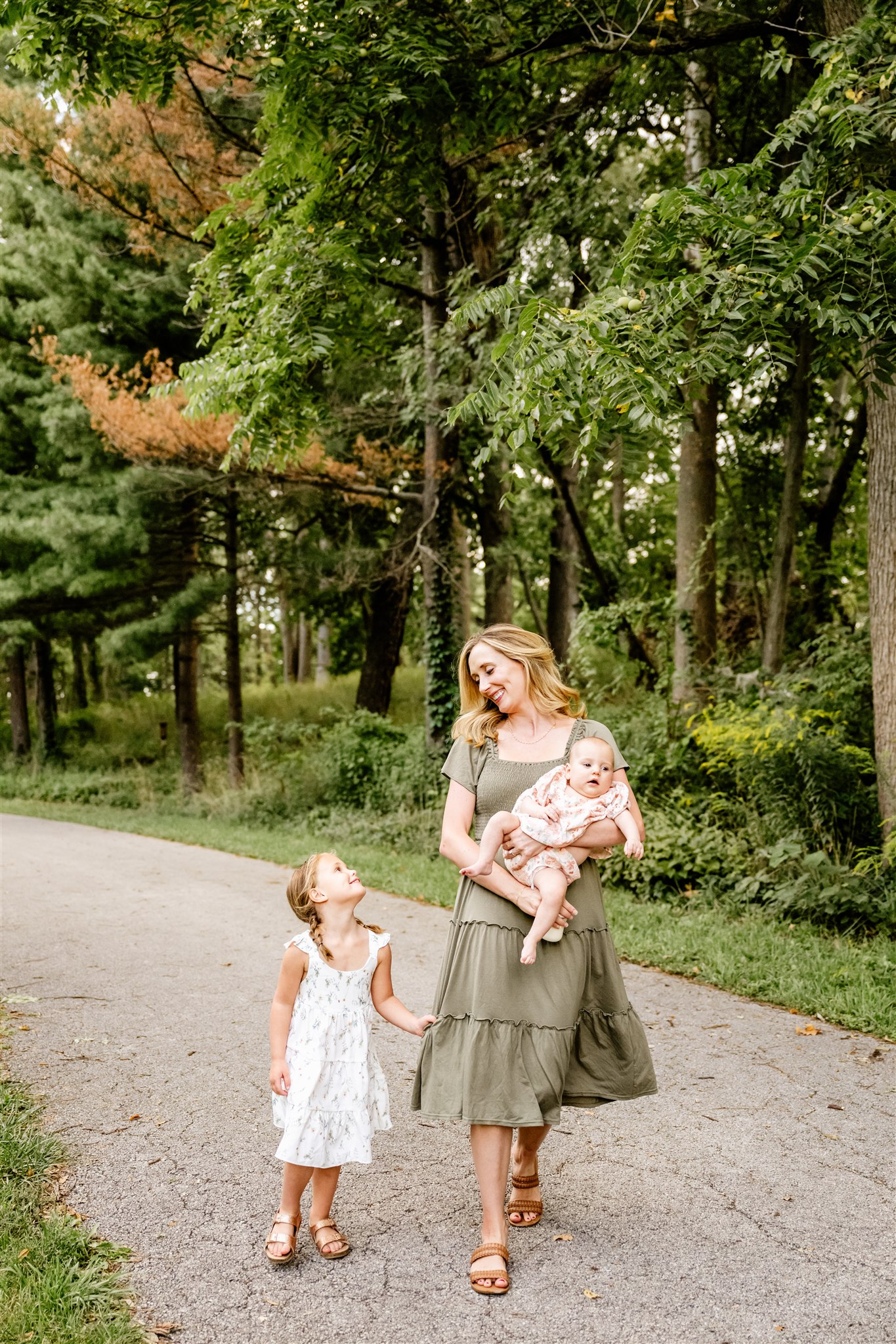 A mom in a green dress holds her baby while her toddler daughter wearing a floral dress clings onto her dress as they walk through a park after visiting splash pads in chicago.