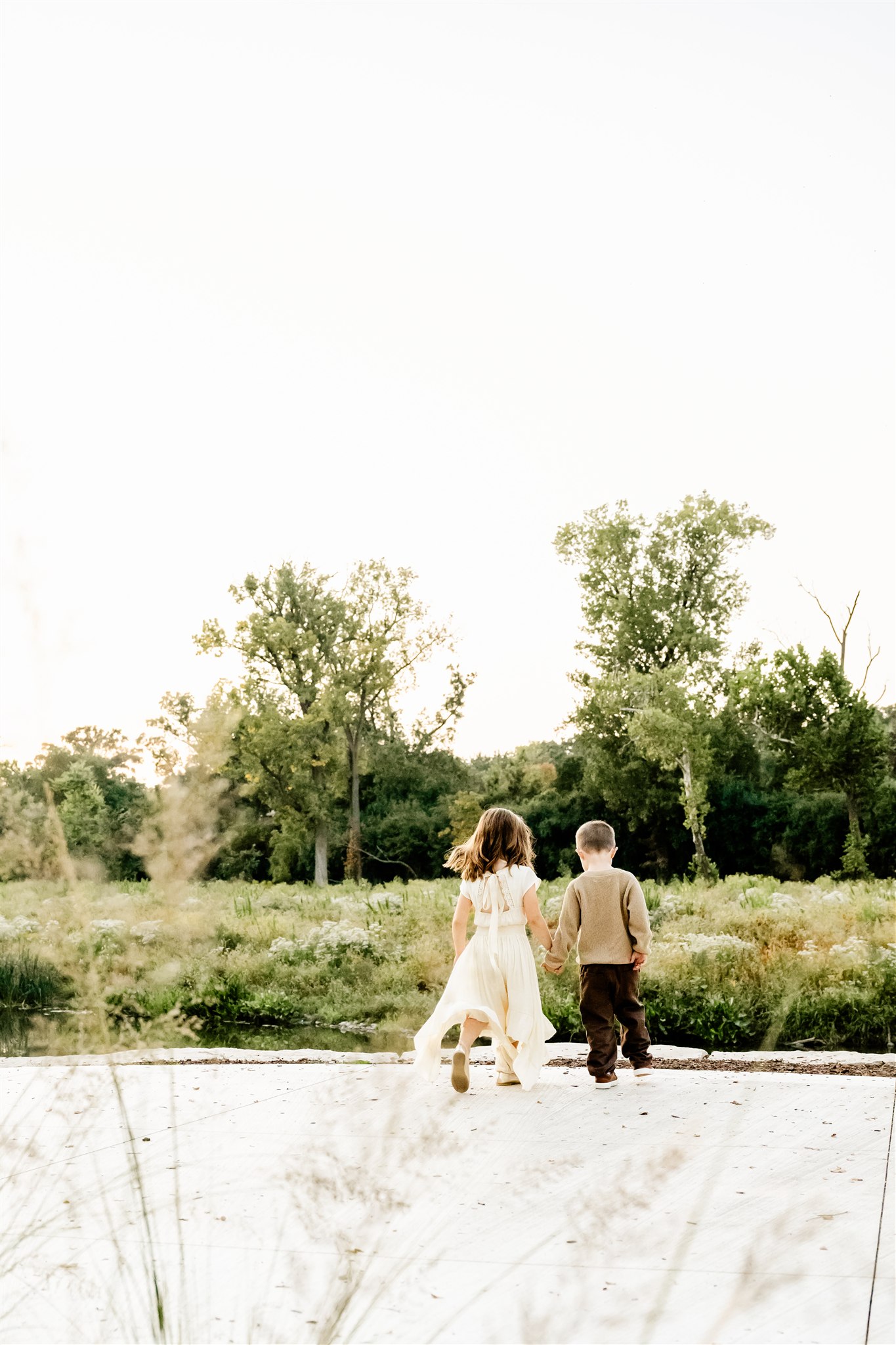 Toddler brother and sister walk holding hands on a park sidewalk exploring a pond
