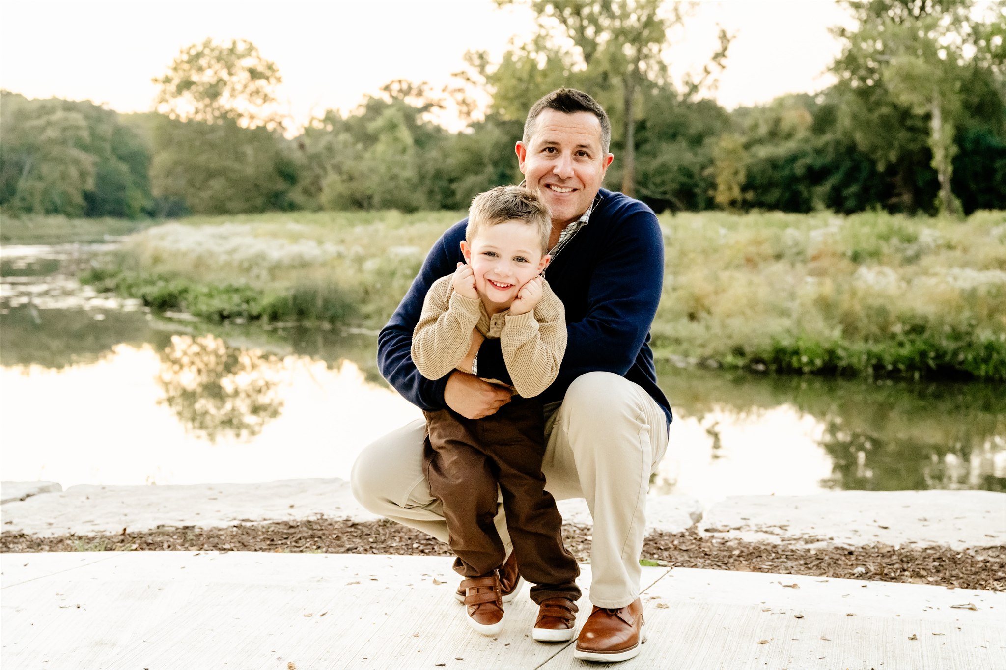 A happy dad in a blue sweater places with his toddler son in a park path by a pond before exploring splash pads in North Shore