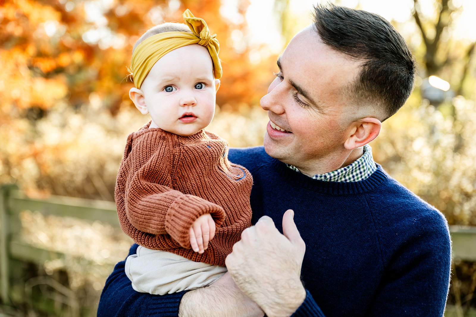 A dad plays with his baby girl in a brown sweater and yellow headband at sunset