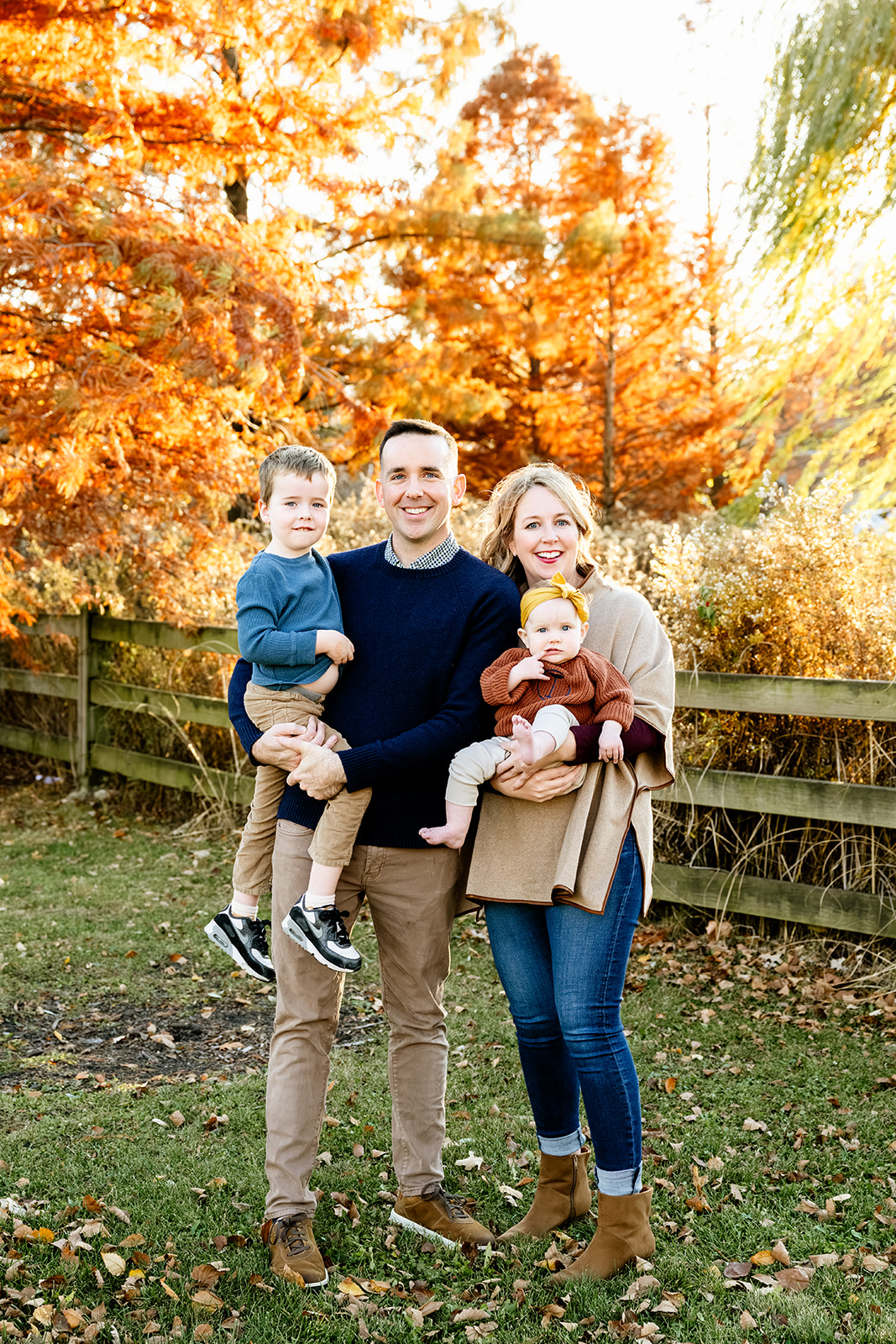 A mom and dad smile while holding their toddler son and daughter in their arms in a park in fall at sunset after visiting baby stores in North Shore Chicago