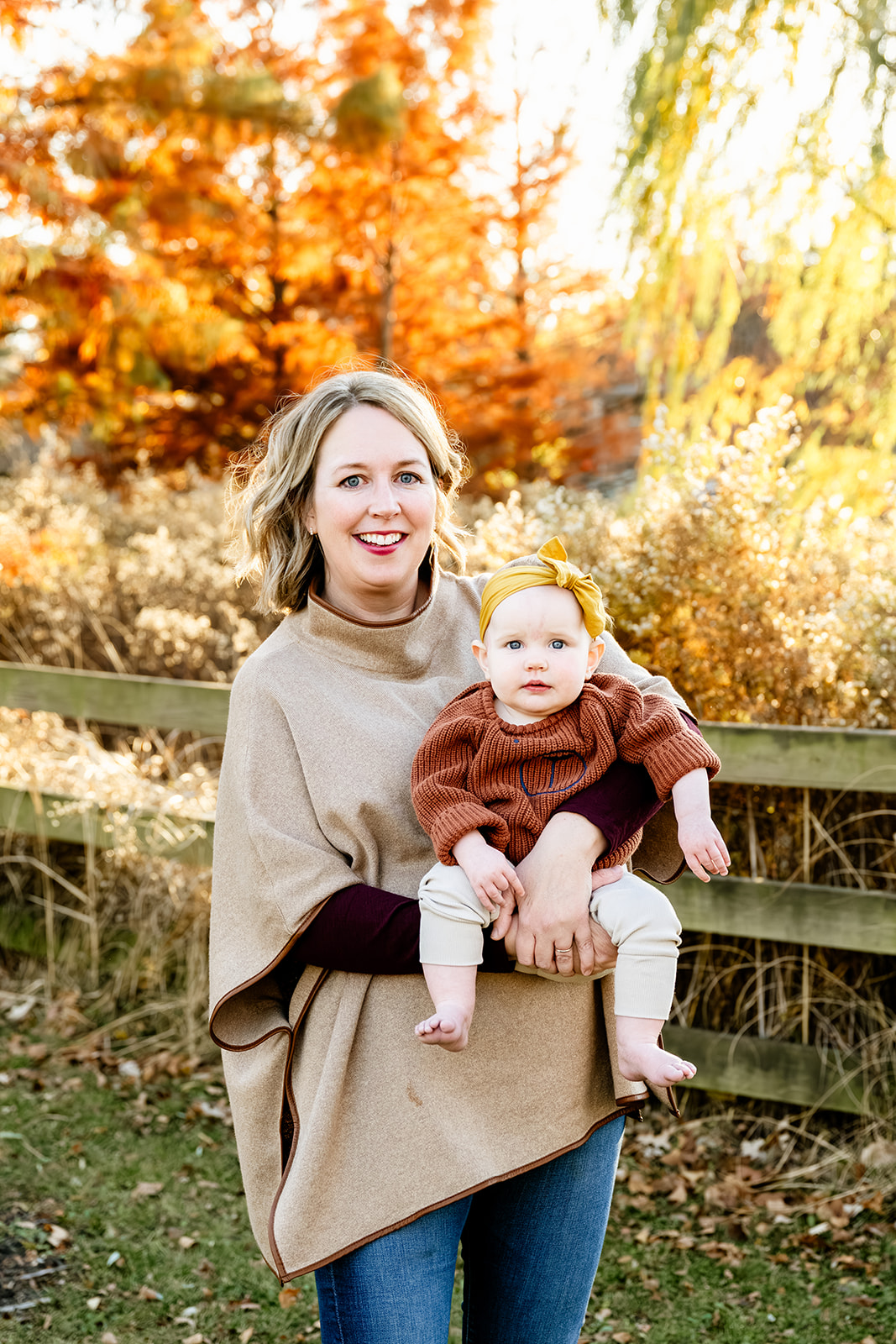 A mother in a brown sweater stands holding her baby girl in a brown sweater in a park at sunset after visiting baby stores in North Shore Chicago