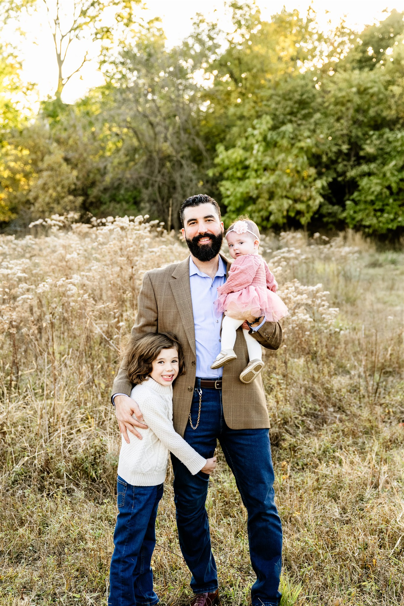 A happy dad stands in a park trail in a tan coat holding his baby daughter and toddler son hugging his leg after visiting childrens boutiques in North Shore Chicago