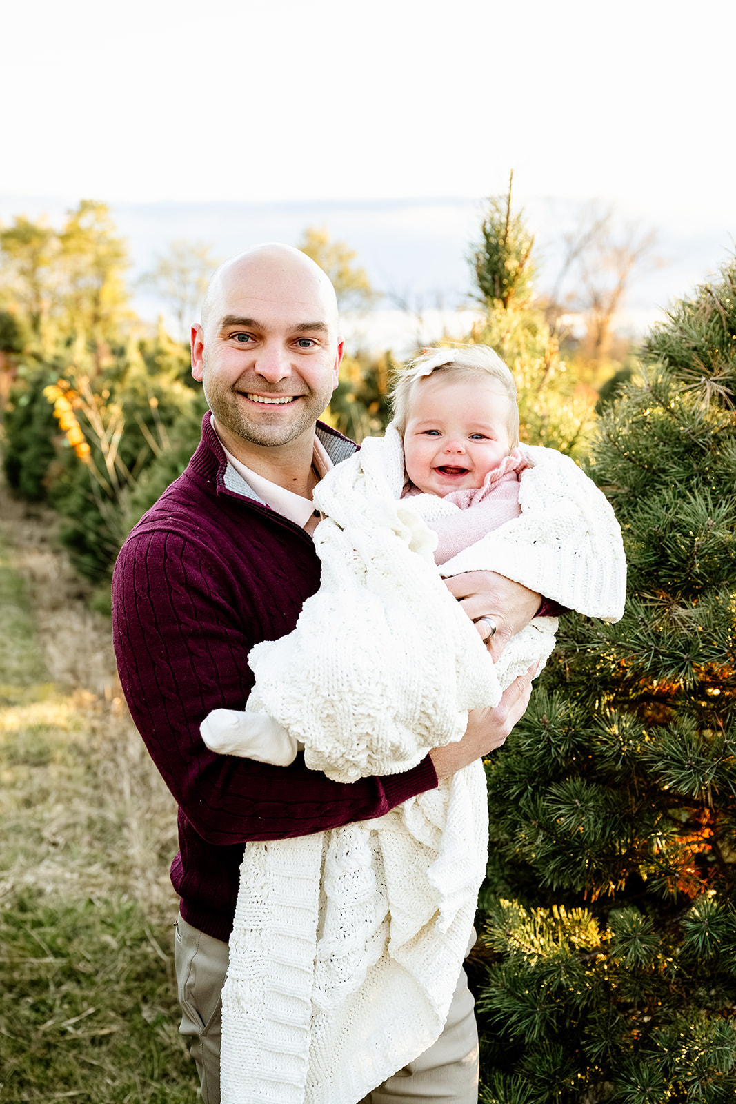 A smiling baby girl in a white blanket sits in dad's arms in a christmas tree farm at sunset ina. pink onesie