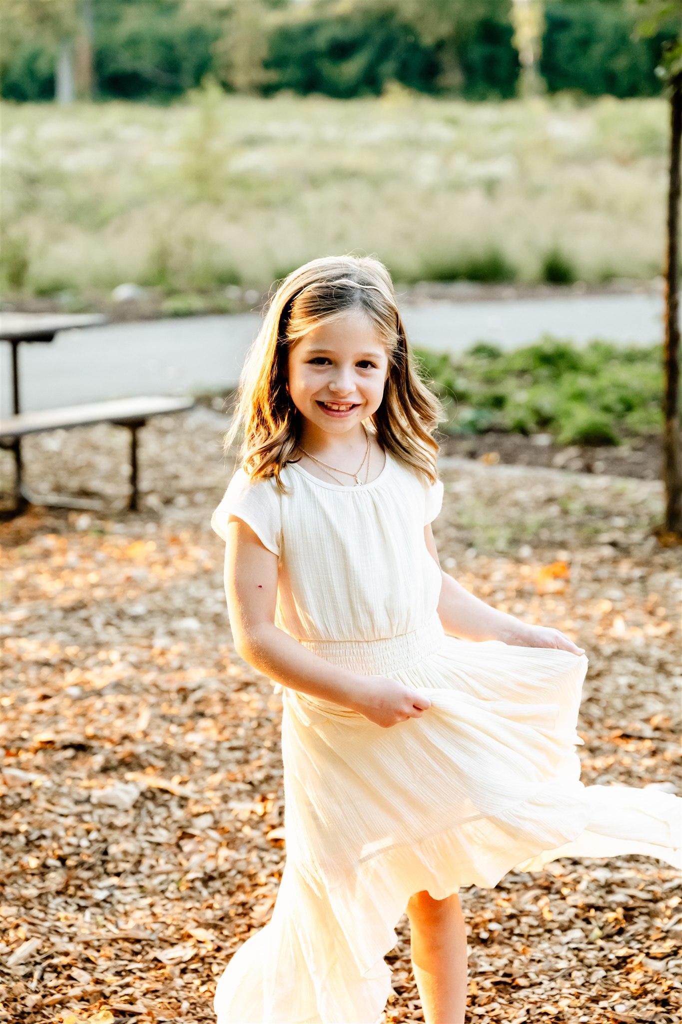 A toddler girl twirls in her cream dress in a park at sunset
