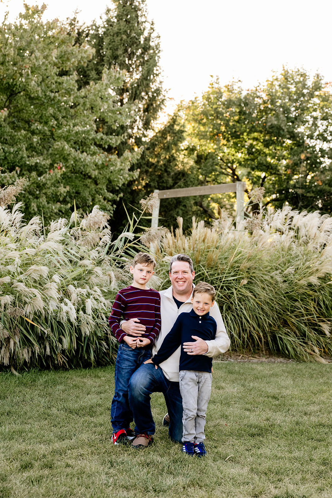 A proud dad kneels in a lawn at sunset hugging his two young sons