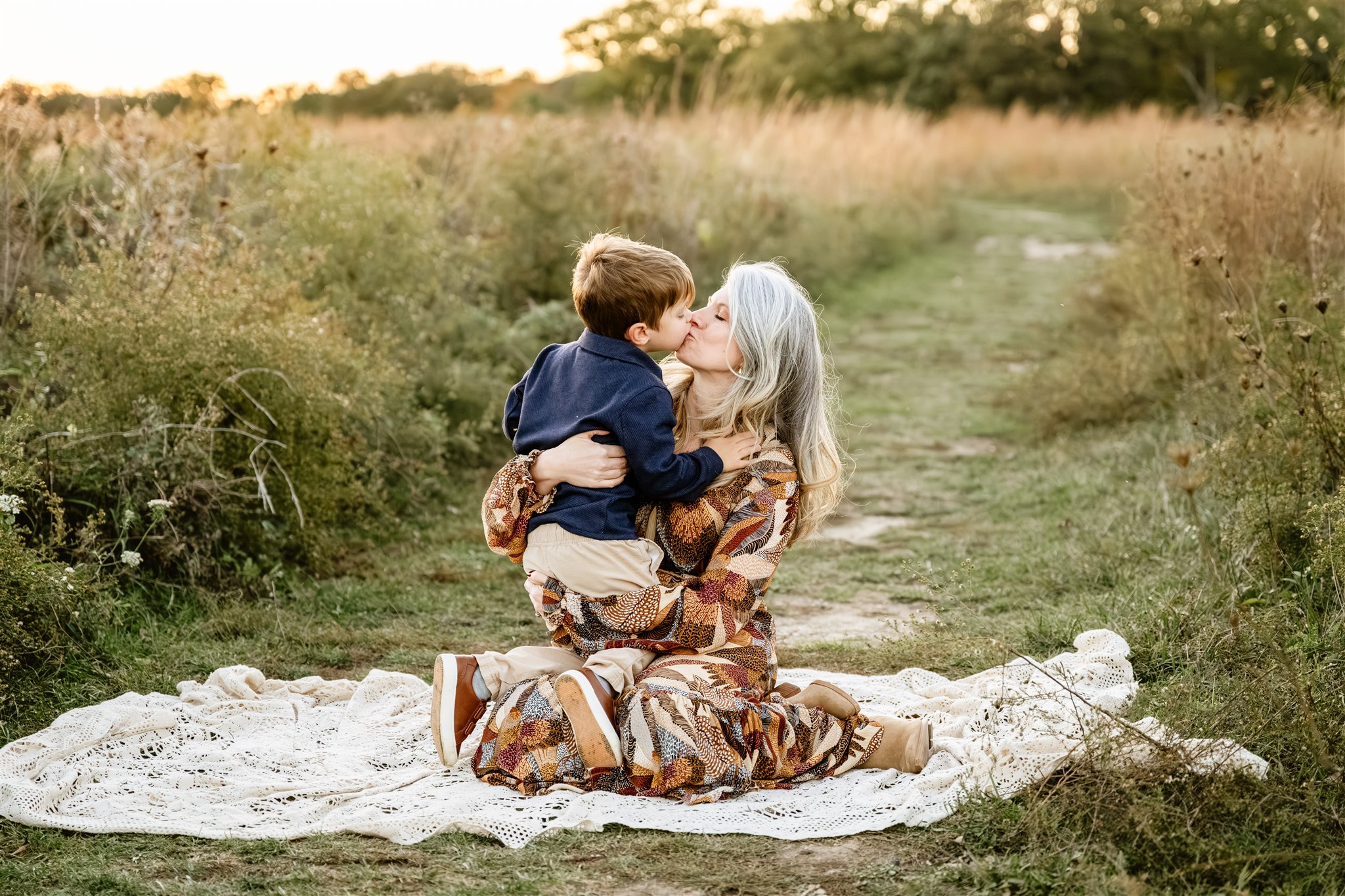 A happy mom in a colorful dress kisses her toddler son while hugging him and sitting on a picnic blanket in a park trail at sunset