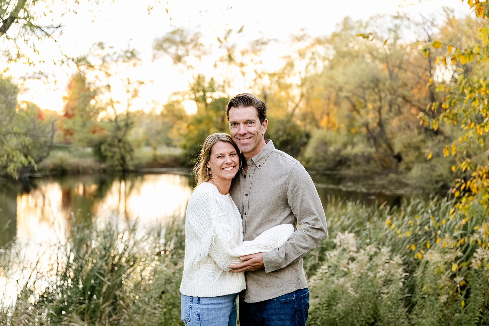 A happy couple stand smiling and hugging by a pond at sunset