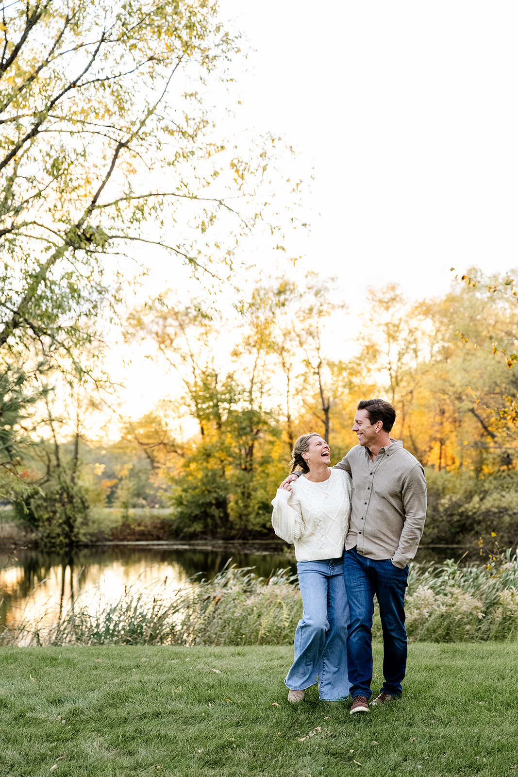 A laughing woman stands with her husband by a pond in cream and tan at sunset before visiting romantic restaurants in North Shore