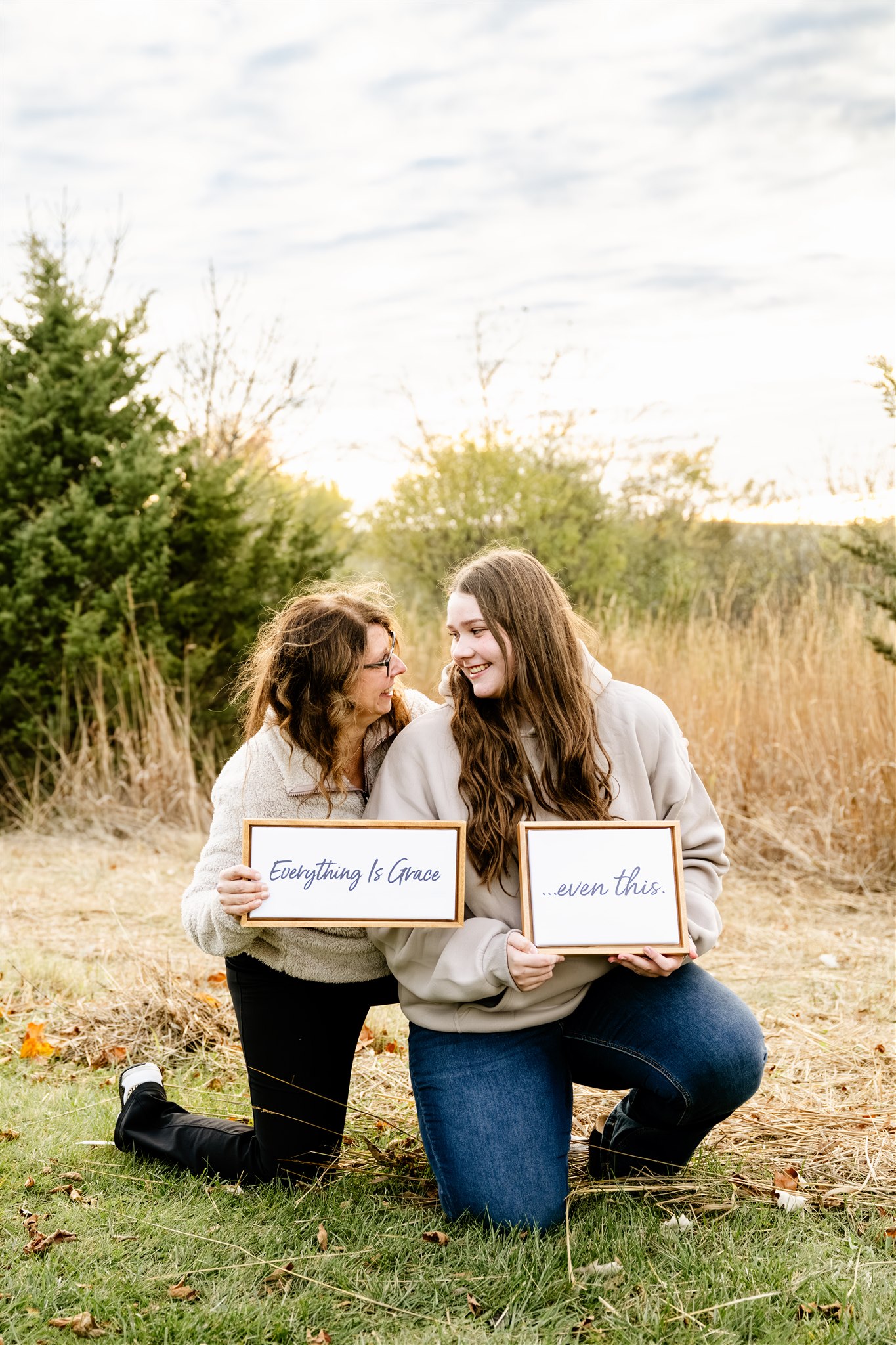 A mom and daughter smile at each other while kneeling in a park trail in matching sweaters holding signs after visiting Chrysalis Custom Hair