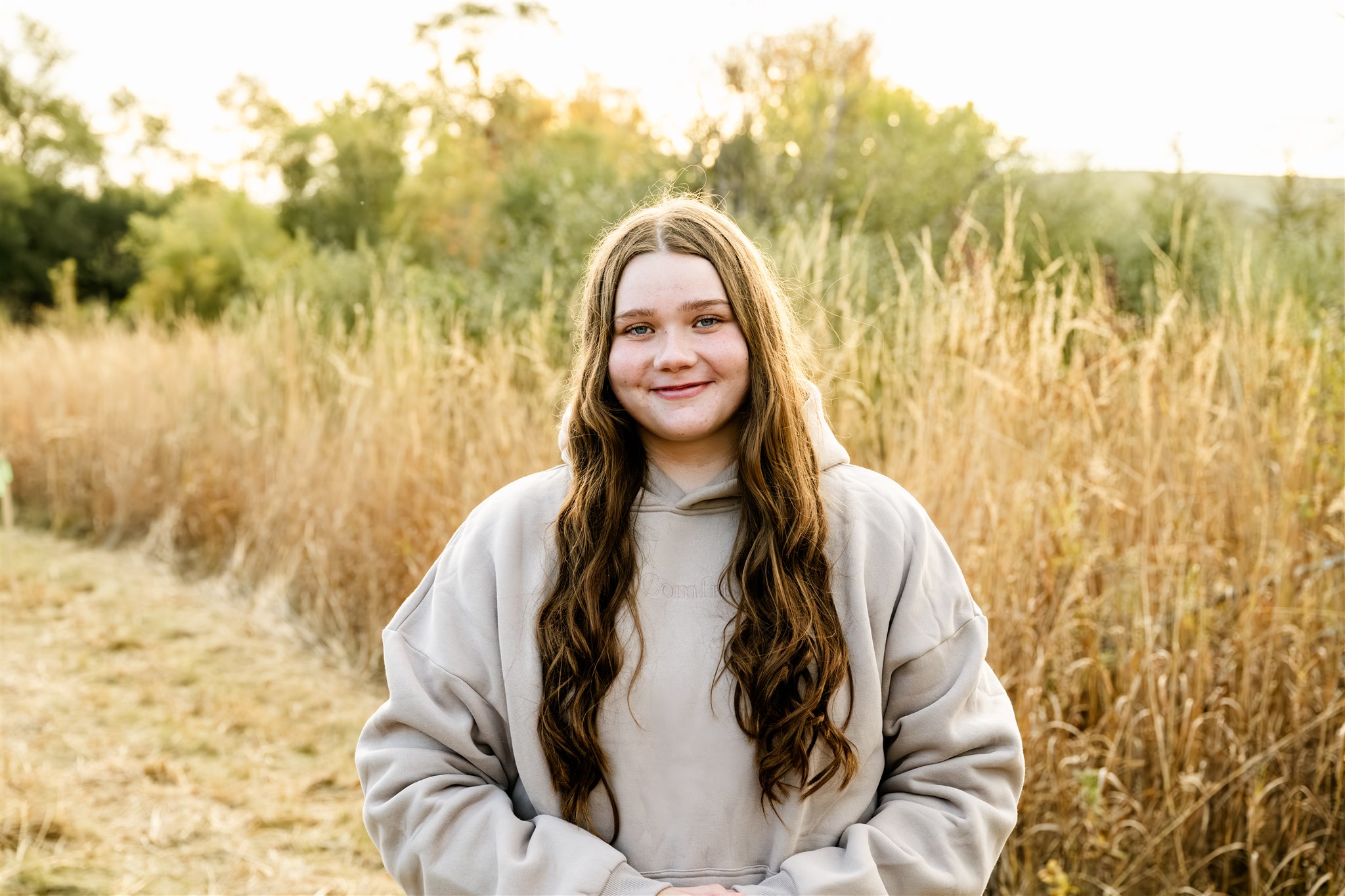 A happy high school senior in a tan hoodie stands smiling in a park trail at sunset after visiting Chrysalis Custom Hair