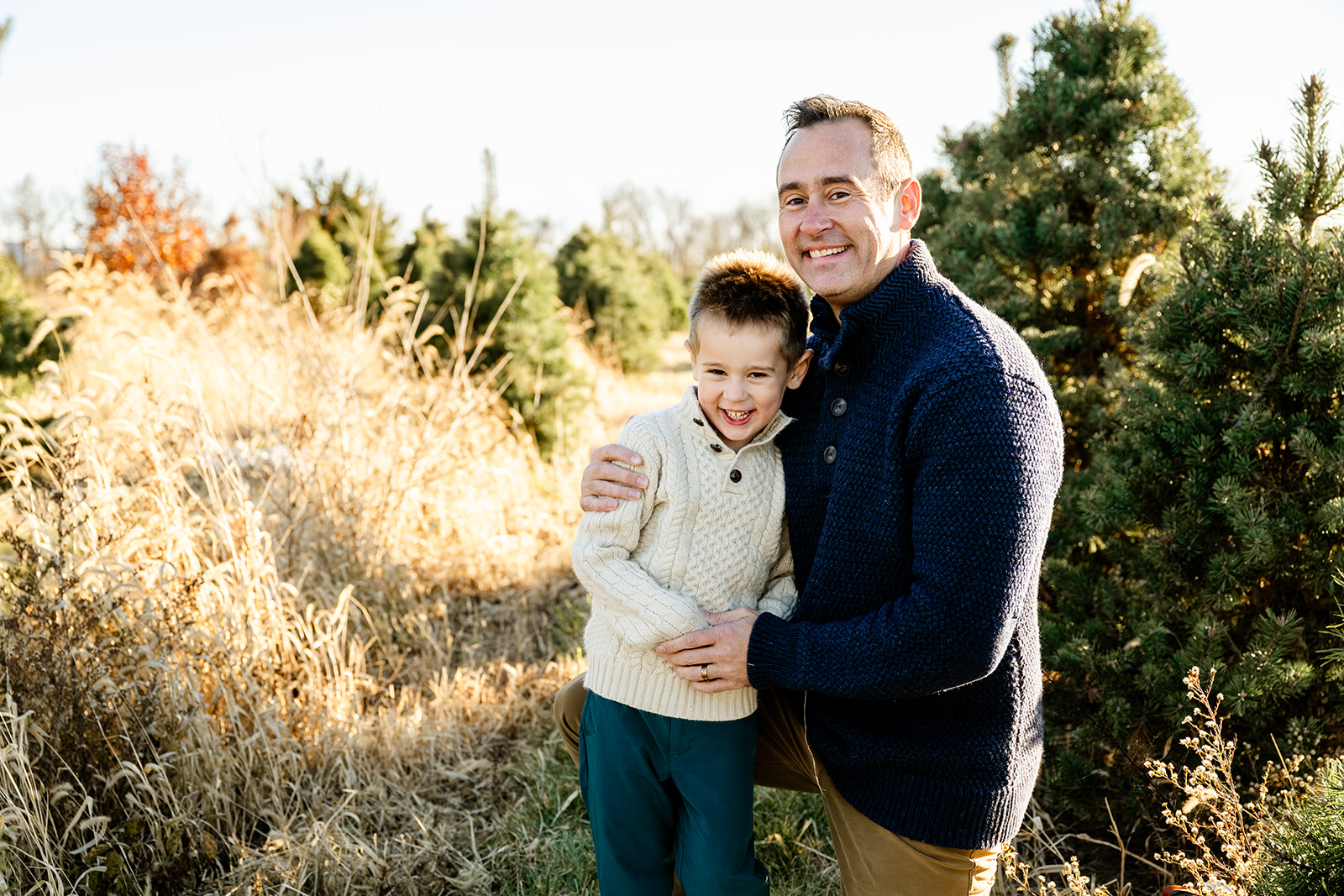 A happy dad in a blue sweater hugs his giggling young son in a cream sweater in a christmas tree farm after visiting Kohl Children's Museum