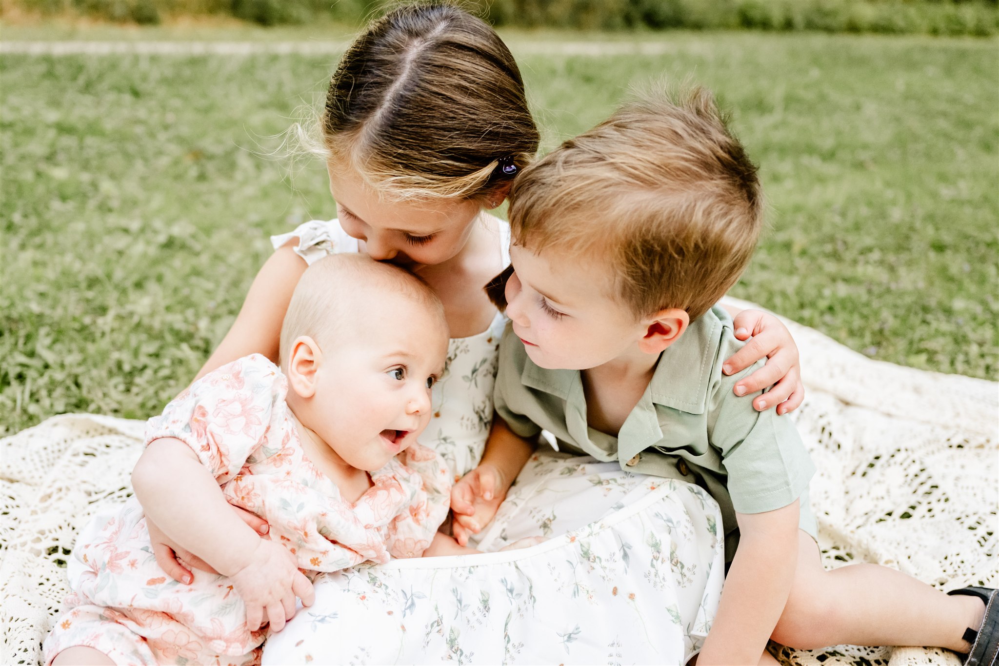 Toddler brother and sister in green and florals cuddle with their their baby sister on a lace picnic blanket