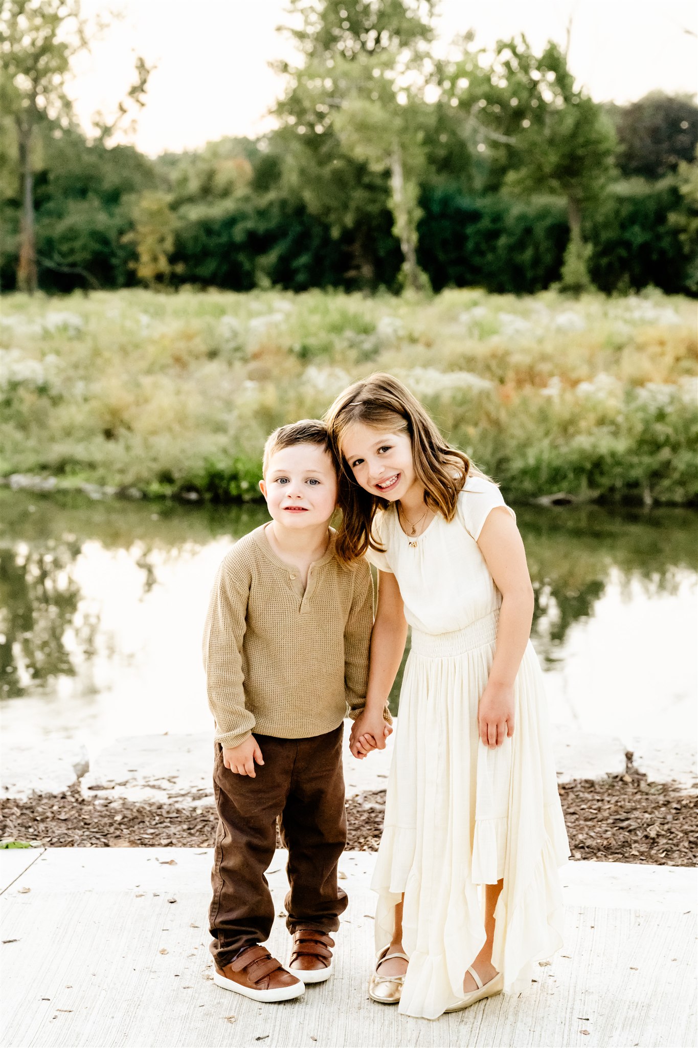 Happy young brother and sister in a white dress and tan sweater stand on a sidewalk holding hands and smiling after visiting a children's ministry in Naperville