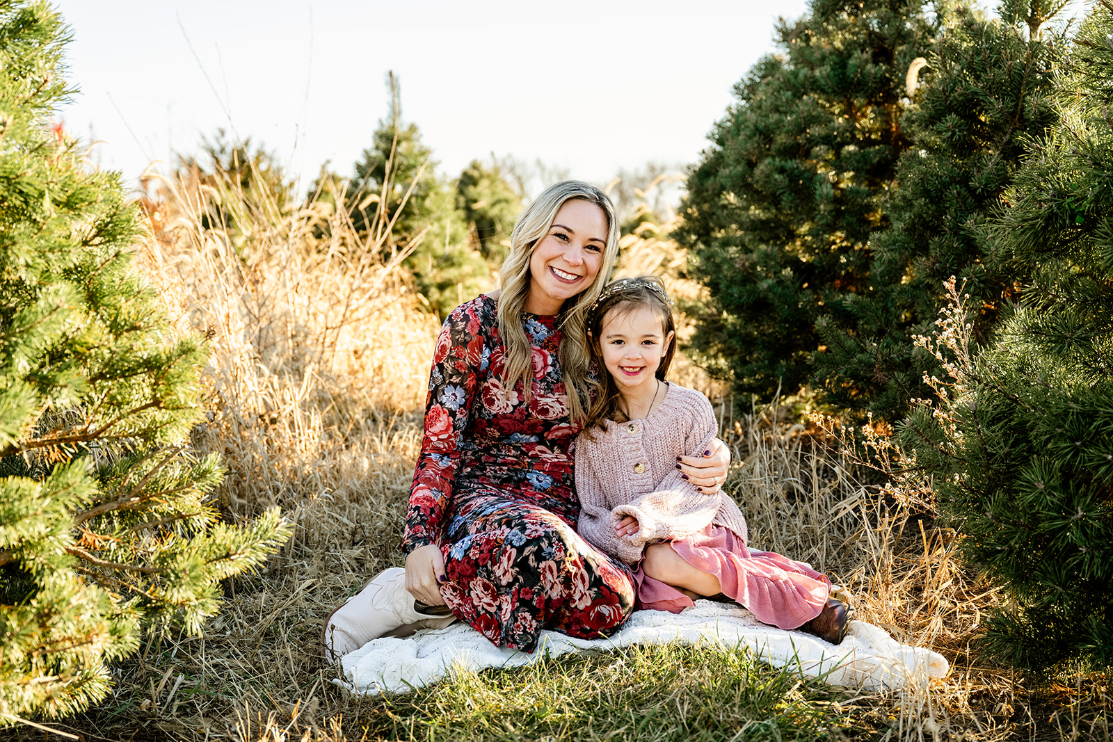 A smiling mother in a floral print dress cuddles her young toddler in a pink sweater among christmas trees after some ice skating in Naperville