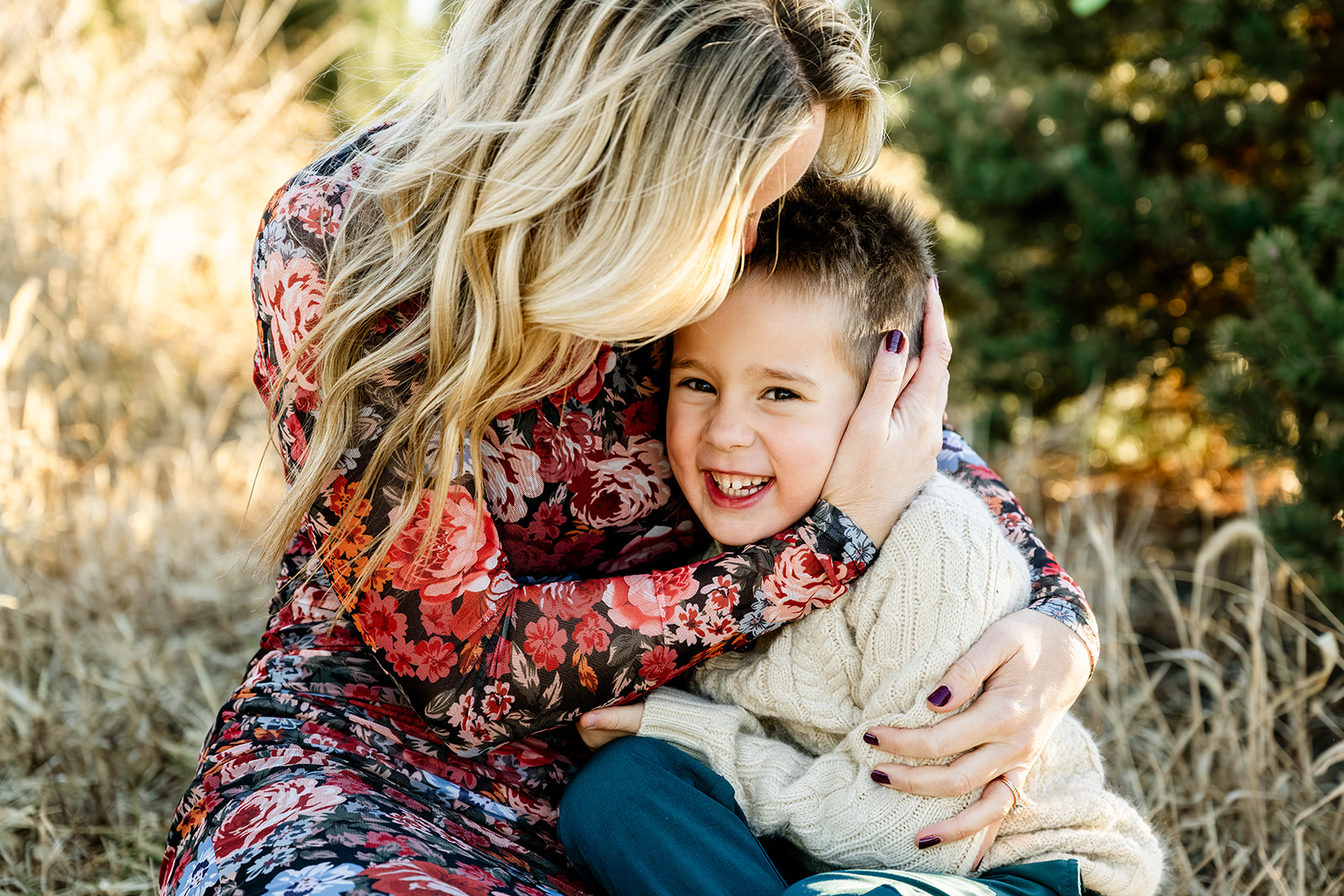 A mother in a floral print dress snuggles her giggling toddler son in a tan sweater in a field of tall golden grass before some ice skating in Naperville